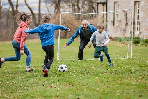 A father playing football with his three children on a grass area while on a staycation in Northumberland. Two children are trying to kick the ball into goal while the father and other child defend the goal.