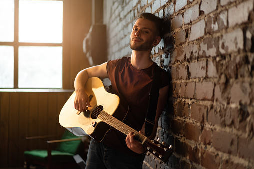 Man with acoustic guitar leaning against brick wall playing music singing songs enjoy life Handsome caucasian male guitar player practice play musical instrument home loft interior Creative lifestyle