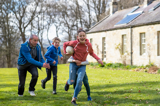 A father playing a game of rugby with his three children in a back yard while they're on a staycation in Northumberland. One boy has his arms around his sister, who is holding the rugby ball. He is trying to tackle her to the ground while her father and other brother is running after them.
