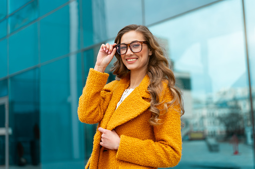 Business woman dressed yellow coat standing outdoors corporative building background Caucasian female business person eyeglasses on city street near office building big windows Stylish businesswoman