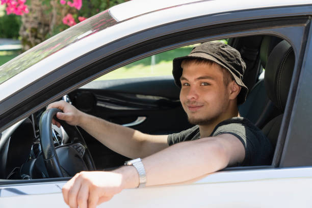 a teenager is driving. a modern young guy, fashionable with a beard, drives a white car. driving training or car travel. a man looks at the camera, portrait stock photo