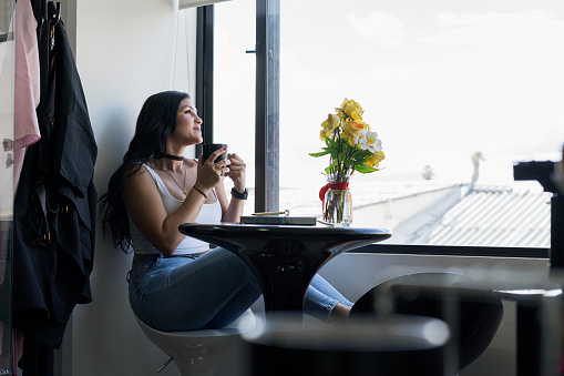 Beautiful Latin woman from Bogota Colombia between 20 and 29 sitting in the window of her house enjoying her morning coffee, while looking and enjoying the landscape