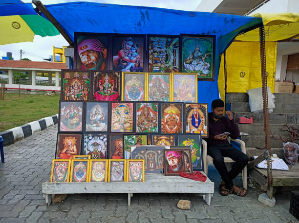 uma loja à beira da estrada vendendo fotos emolduradas de deuses hindus e deusas e o vendedor absorto em navegar em seu smartphone em mysuru, índia. - shiv bangalore shiva god - fotografias e filmes do acervo