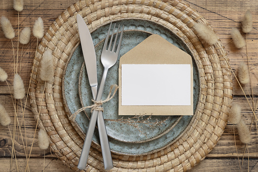 Table place with blank paper card, envelope, fork and knife on plates surrounded by dried flowers top view on wooden table. Boho wedding card mockup