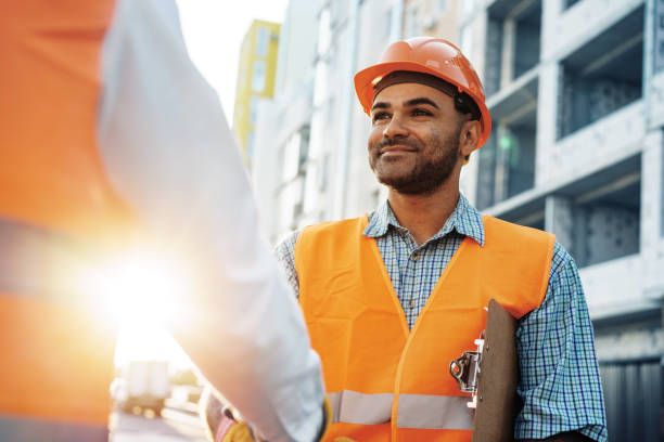 Two men engineers in workwear shaking hands against construction site.