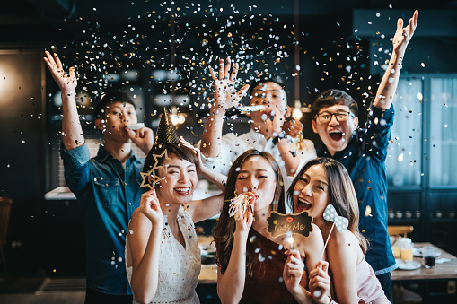 A young group of cheerful Asian man and woman having fun with props and blowing sparkling confetti at party