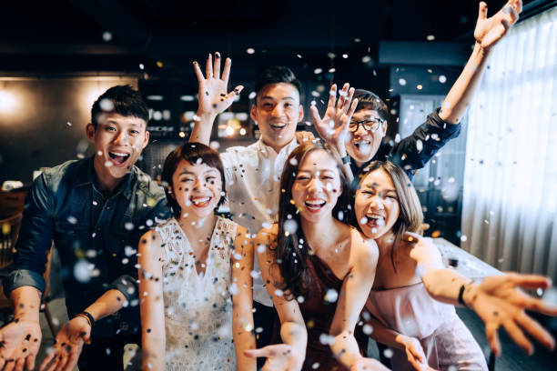 A young group of cheerful Asian man and woman having fun and blowing sparkling confetti at party stock photo