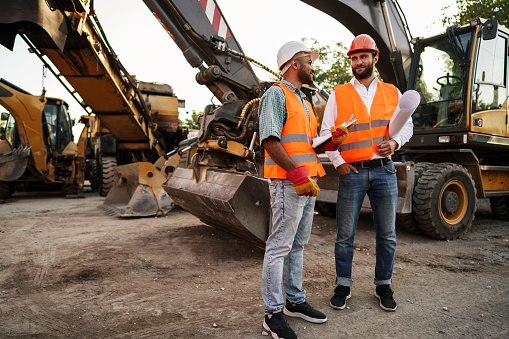 Two men engineers in workwear discussing their work standing against construction machines