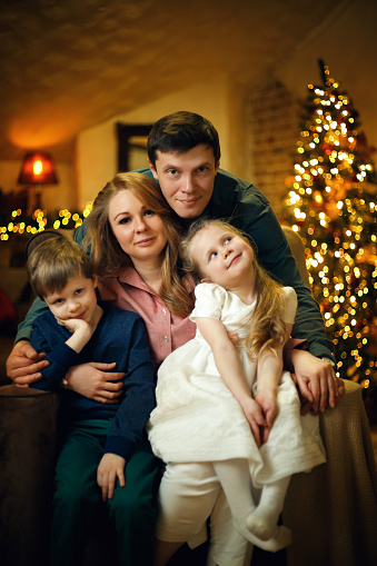 Young beautiful caucasian family with two children posing in a chair in a Christmas interior with a christmas tree and garlands. Selective soft focus, film grain effect