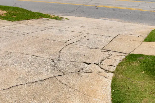 Photo of Cracked and broken aggregate concrete driveway descending into an asphalt roadway, flanked by green grassy areas, creative copy space