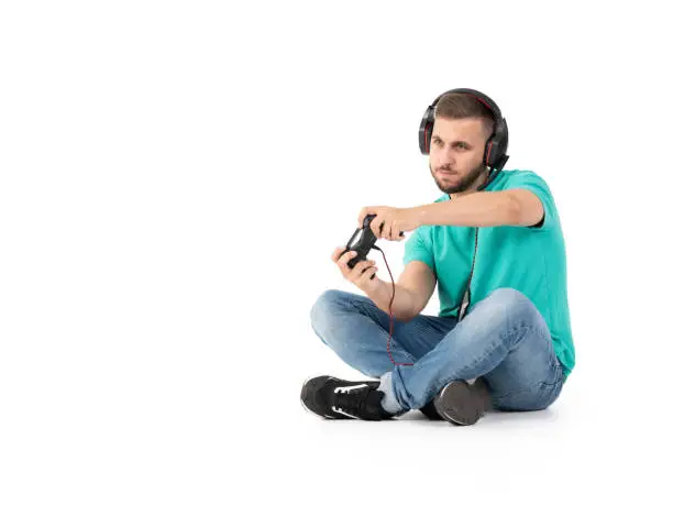 Young man playing videogames with a controler and a headphones in a white background with jeans, black shoes and green shirt.