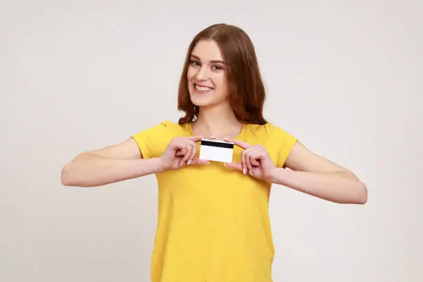 Photo of Portrait of happy beautiful young woman in yellow casual style T-shirt showing credit card, looking at camera with toothy smile, banking.