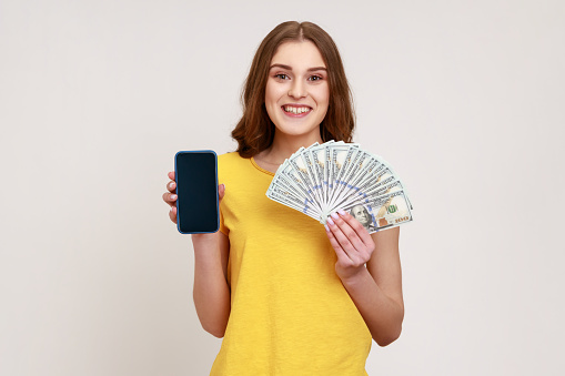 Portrait of successful happy young woman with pleasant smile wearing casual style T-shirt holding money and phone, looking at camera. Indoor studio shot isolated on gray background.