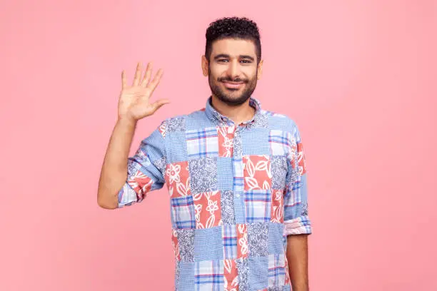 Hello, welcome! Portrait of happy young adult dark haired bearded man in blue shirt raising palm to wave hi, greeting with hospitable friendly smile Indoor studio shot isolated on pink background.