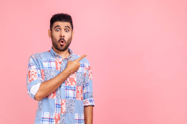 istock Excited surprised handsome man with beard wearing blue casual shirt looking at camera with open mouth, pointing finger aside, showing copy space. 1343293402