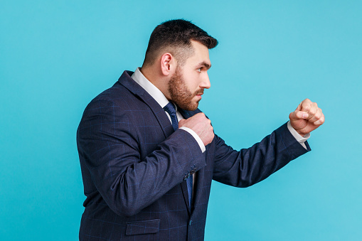 Side view of aggressive businessman wearing official style suit being ready to punch, boxing with clenched fists, fighting with somebody. Indoor studio shot isolated on blue background.