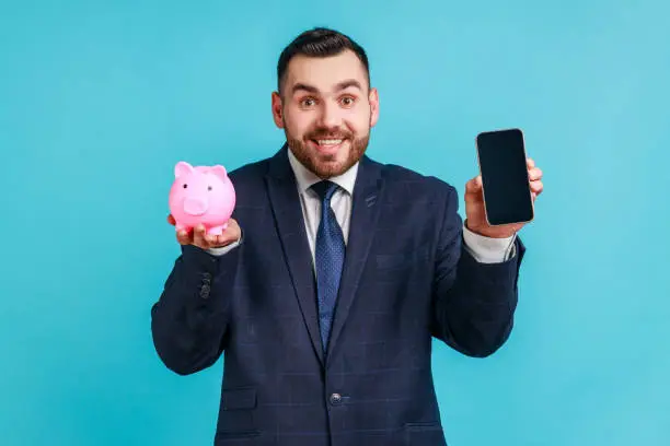 Photo of Happy smiling man with beard wearing official style suit holding smartphone with empty screen and piggy bank, cashback, online wallet.