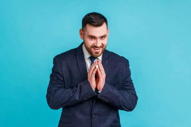 Portrait of devious handsome brunette man with beard wearing dark official style suit, thinking devious tricks and cheats, looking at camera. Indoor studio shot isolated on blue background.