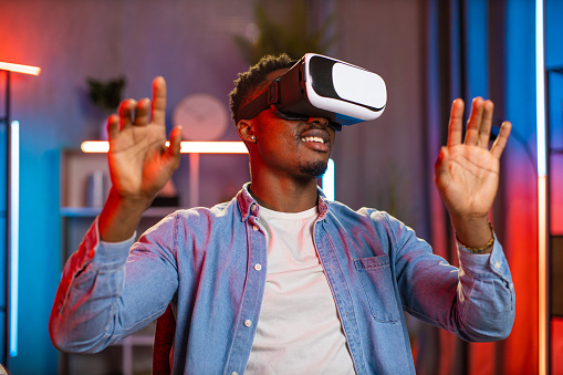 Likable smiling 30-aged African American man in virtual 3d glasses, working on imaginary display in the evening in home office. Man playing video games at home using vr goggles
