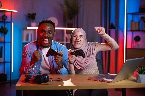 Joyful African man and Muslim woman feeling happiness while winning in video games on laptop. Multi ethnic couple enjoying entertainment at home during evening time.