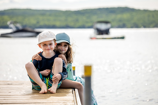 A cute redhead boy and a cute little girl are sitting on a pier. The brother and sister are embracing and looking at the camera and smiling. They are spending their summer vacations at the lake and beach.