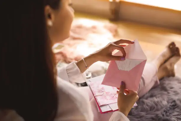 Selective focus shot of young woman sitting on the bedroom floor and opening a love letter.