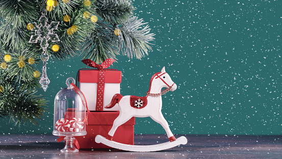 Christmas ornaments and gift boxes on green with snow. Rocking horse under Christmas tree. New Year greeting card. Copy space banner