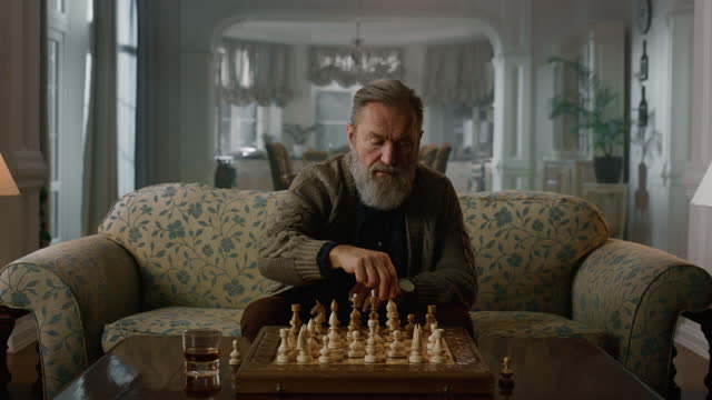 Senior man playing chess alone in classic living room. Focused mature gentleman