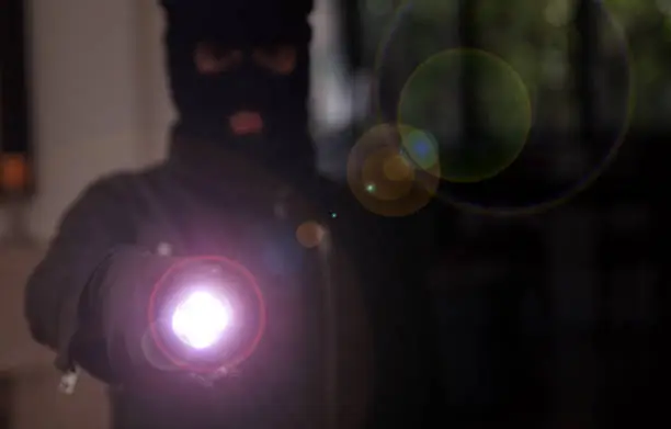 Housebreaking, burglary concept. Masked burglar in black balaclava entering a house holding a flashlight, dark night background.