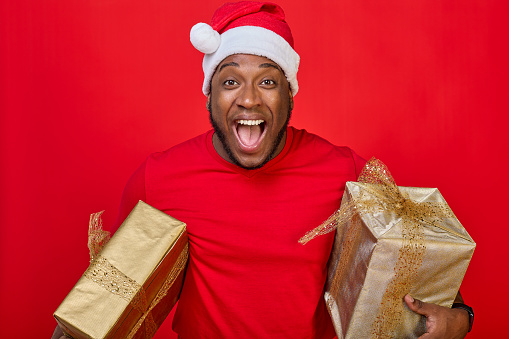 Portrait of a black guy with a toothy smile in a Santa hat holding Christmas gifts under his armpits