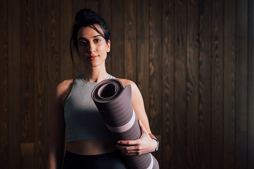 Young female in sportswear looking at camera while standing with exercise mat in hand over a dark wooden wall