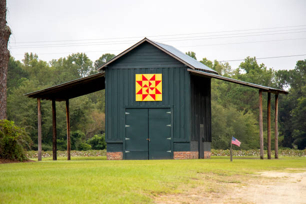 A bright windmill pattern quilt on a painted wooden pole barn in rural Georgia, USA Lenox, Georgia - September 17, 2021: A painted wooden pole barn on a rural Georgia cotton farm displays a traditional windmill quilt pattern in red and yellow. A cotton field is visible in the background. patriotic quilt stock pictures, royalty-free photos & images