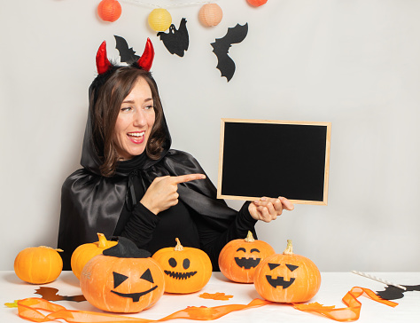 Young woman wearing in a witch costume holding blank empty banner smiling happy pointing points the finger to the place for the text.