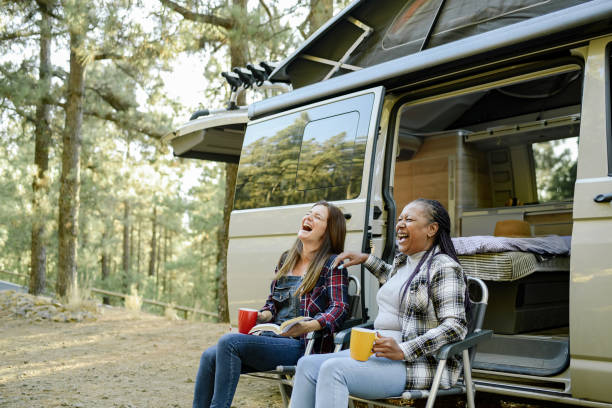 Multiracial traveling women laughing near camper Excited diverse female friends sitting in camping chairs near traveling van and laughing while having fun in woods mobile home stock pictures, royalty-free photos & images