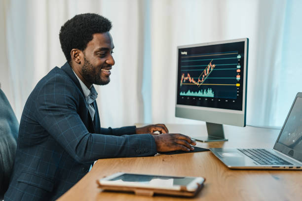 Black man working with binary option Side view of smiling male trader in formal suit using laptop and computer monitor with binary option charts while working at table wages photos stock pictures, royalty-free photos & images