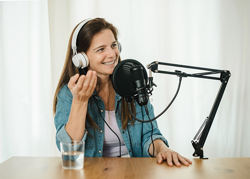 Smiling female in headphones recording podcast and speaking in modern mic while sitting at table and gesticulating