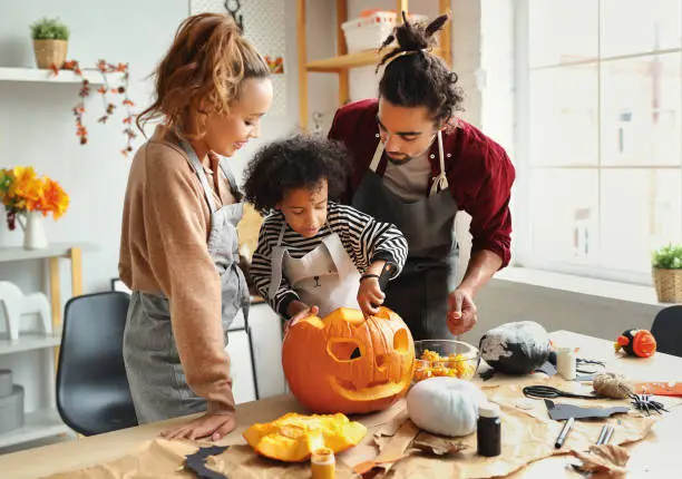 Happy ethnic family mother, father and son carving pumpkin for Halloween holiday together, preparing for holiday party in kitchen,  having fun while creating Jack-o-lantern