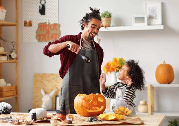 Ethnic father removing pulp from ripe pumpkin while carving jack o lantern with little son for Halloween Smiling african american parent father removing pulp from ripe pumpkin while carving jack o lantern with little son for Halloween celebration at home in kitchen and looking at each other with smile carving food photos stock pictures, royalty-free photos & images