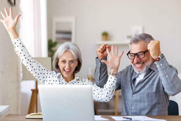 couple de personnes âgées heureux célébrant le succès assis à table avec un ordinateur portable ouvert à la maison - women computer couple surprise photos et images de collection