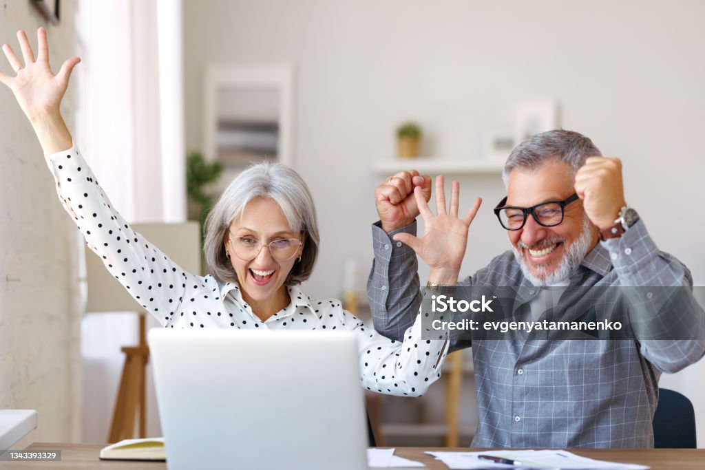 Happy senior couple celebrating success while sitting at table with open laptop at home Happy senior couple celebrating success while sitting at table with open laptop at home and screaming from excitement after getting good news, excited retired family winning lottery Ecstatic Stock Photo