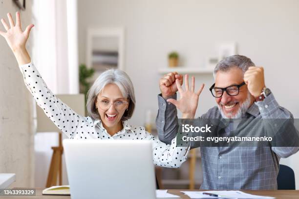 Happy Senior Couple Celebrating Success While Sitting At Table With Open Laptop At Home Stock Photo - Download Image Now
