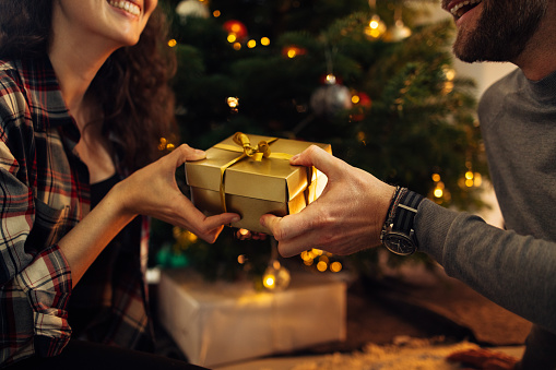 Close-up of man and woman holding a Christmas gift box in hand at home. Couple exchanging Christmas gifts.