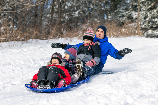 A family is sledding on the snow outdoors in winter. The father and his three children, one boy and toy girls, are laughing and having fun together while bonding. It is a beautiful sunny winter day. They could be at the playground or in their backyard.