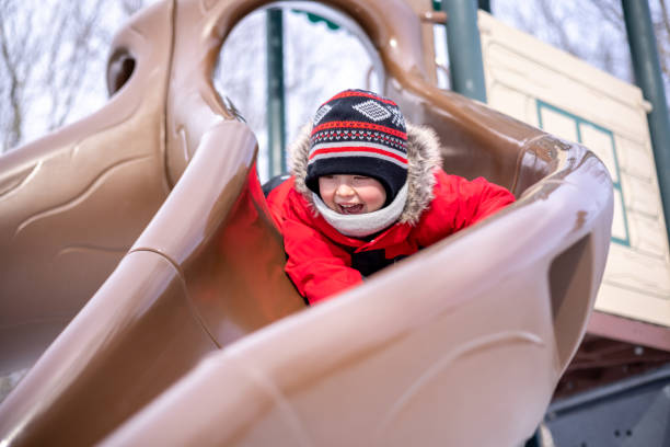 Little Boy Sliding Down a Slide at the Playground Outdoors in Winter. He is very happy and smiling. It is a cold winter day and he is wearing warm clothing.
