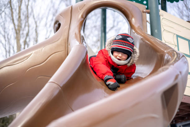 Little Boy Sliding Down a Slide at the Playground Outdoors in Winter. He is very happy and smiling. It is a cold winter day and he is wearing warm clothing.