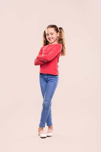 Full body of positive preteen girl with ponytails dressed in casual red sweatshirt and jeans standing with arms crossed and looking at camera