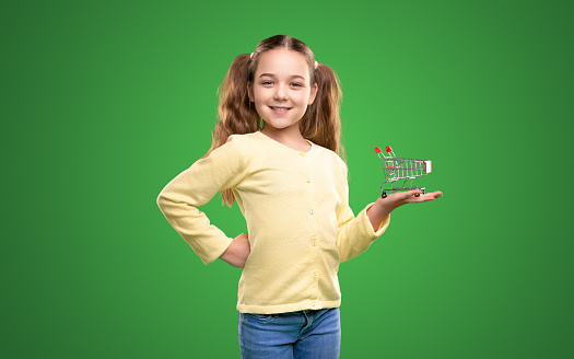 Confident smiling girl in casual clothes showing mini toy shopping cart on hand while representing supermarket against green background