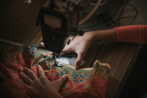 close up top view high angle asian chinese teenage girl's hand holding batik sewing stitching on sewing machine illuminated by led light