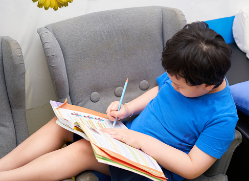 Asian boy sitting on sofa with homework book and use a pencil to do the assigned homework at home