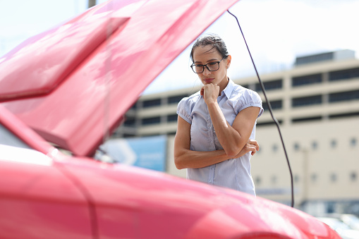 Pensive woman looks at car engine. Car breakdown on the road concept